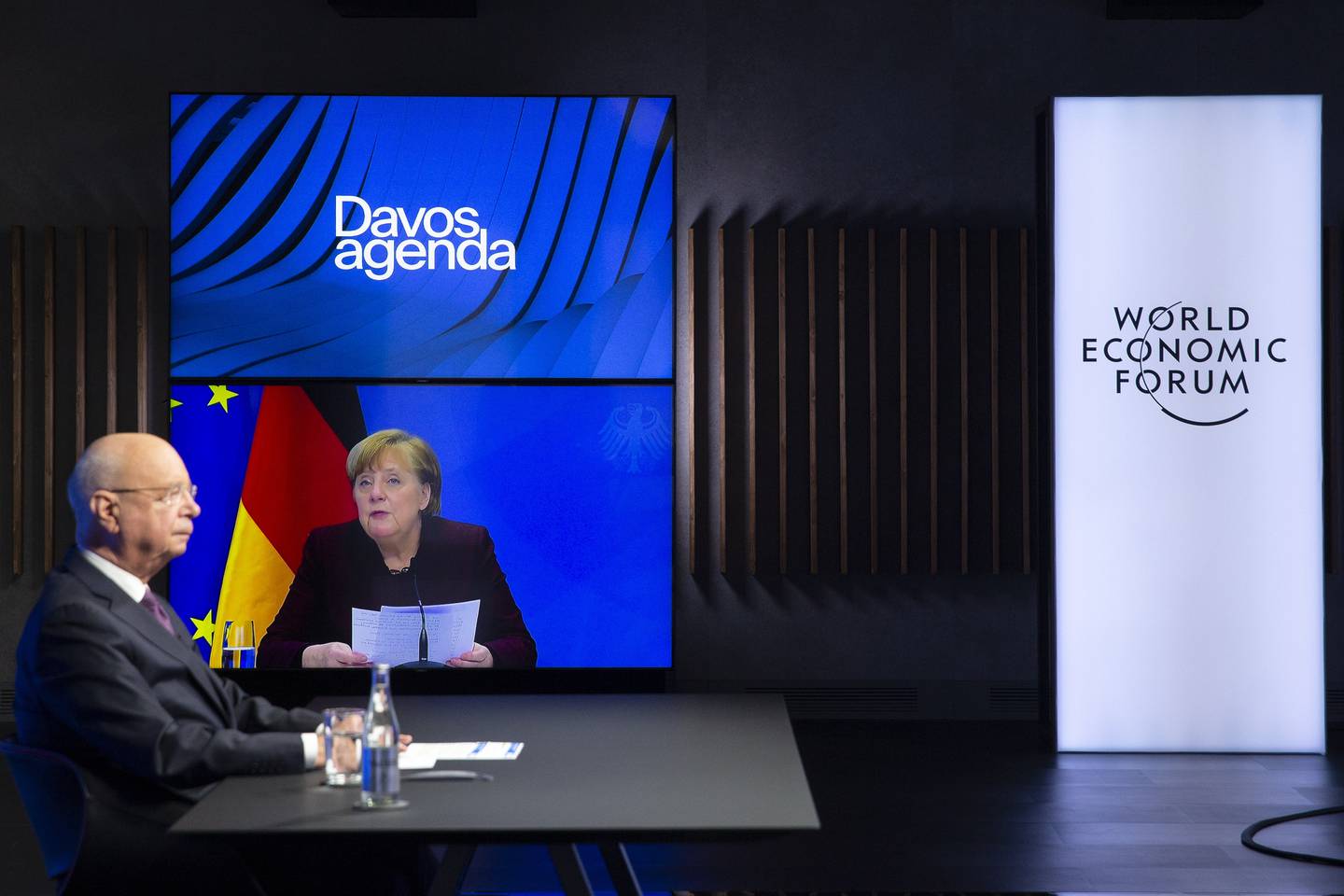 German Klaus Schwab, left, Founder and Executive Chairman of the World Economic Forum, WEF, listens to German Chancellor Angela Merkel, displayed on a video screen, during a conference at the Davos Agenda in Cologny near Geneva, Switzerland, Tuesday, Jan. 26, 2021. The Davos Agenda from Jan. 25 to Jan. 29, 2021 is an online edition due to the coronavirus disease (COVID-19) outbreak. (Salvatore Di Nolfi/Keystone via AP)