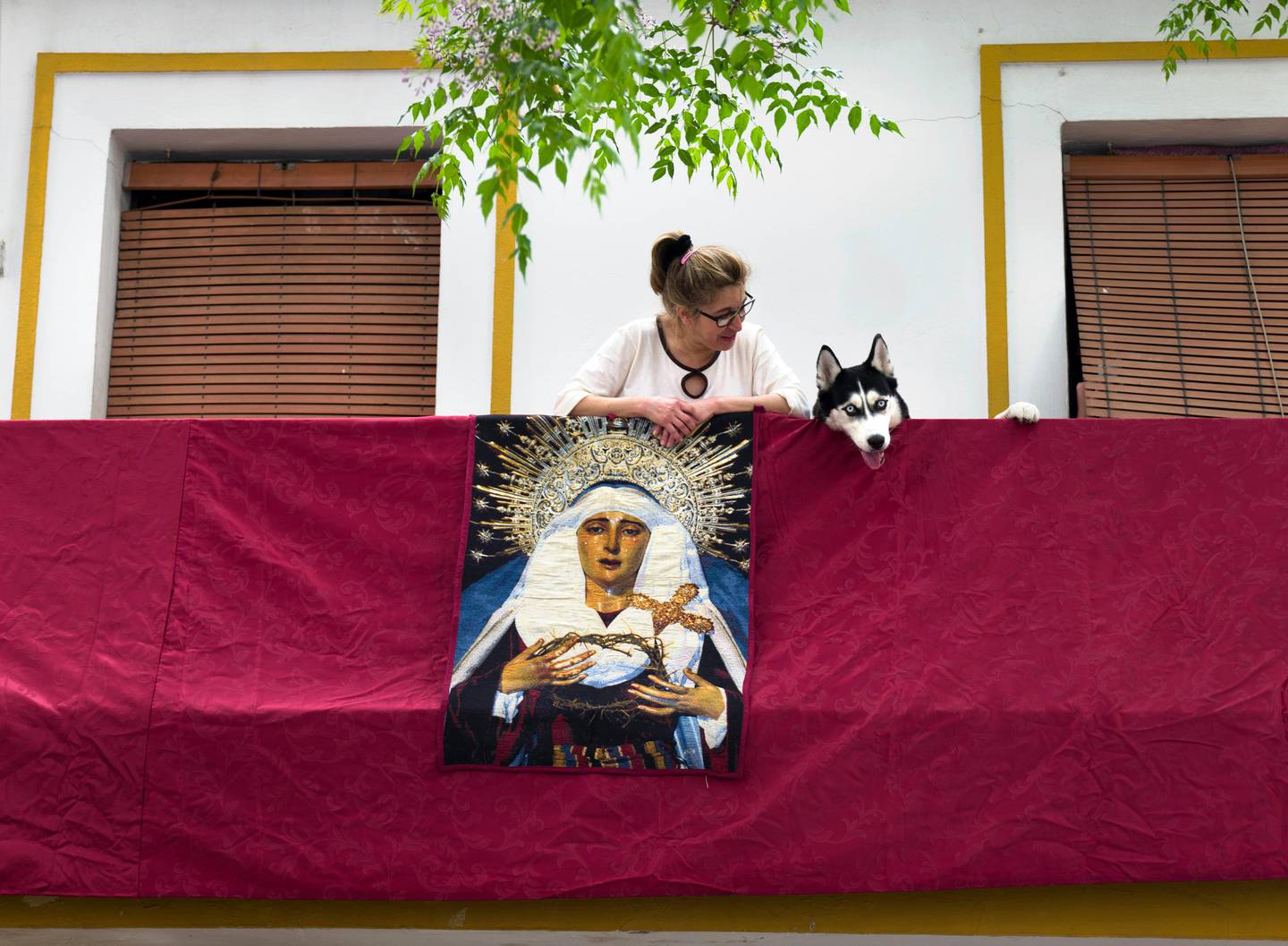 A woman and her dog lean over a balcony with a hanging picture of El Cerro brotherhood's Virgin Mary, after an Easter Holy Week procession was cancelled due to the coronavirus outbreak in Seville, Spain, Tuesday, April 7, 2020. The COVID-19 coronavirus causes mild or moderate symptoms for most people, but for some, especially older adults and people with existing health problems, it can cause more severe illness or death. (AP Photo/Laura Leon)