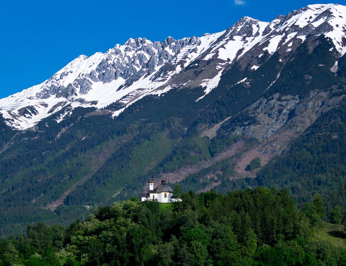 A small chapel stands on a hill in front of the North Chain mountains in Innsbruck, Austria, Saturday, June 1, 2019.  Snow persists on the peaks while lower slopes sport summer flowers with lush green fields. (AP Photo/Michael Probst)