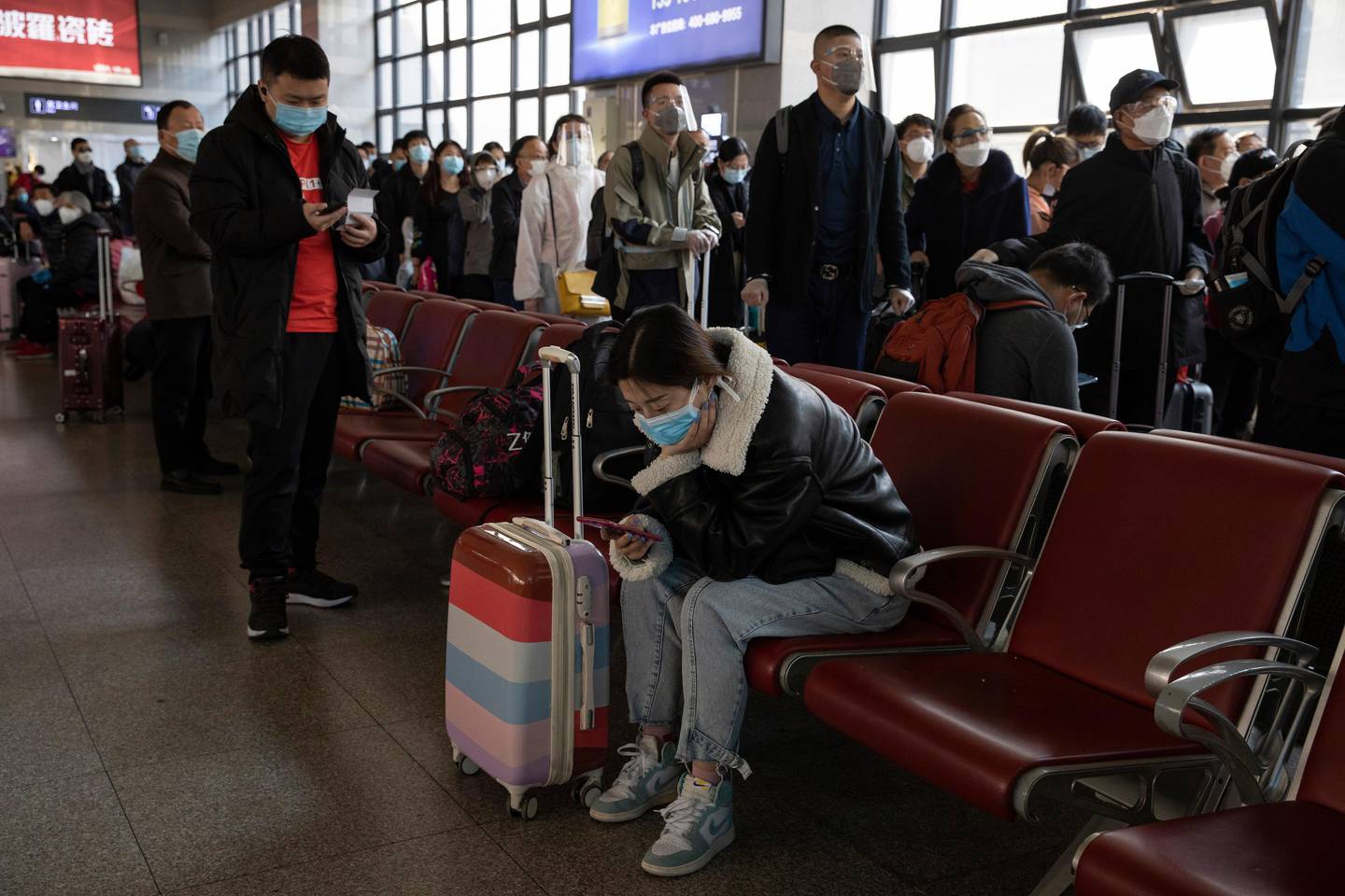 A woman wearing a mask checks her phone as others line up to board a train at a train station in Beijing on Sunday, March 29, 2020. As the coronavirus epicenter has shifted westward, the situation has calmed in China, with falling death rates and most new cases coming from abroad, restrictions on travel have been slowly lifted. (AP Photo/Ng Han Guan)